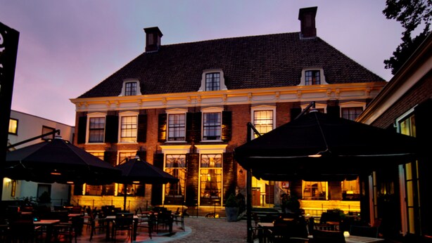Een gebouw met een geplaveide binnenplaats, buitenterras met parasols en een bakstenen gevel.