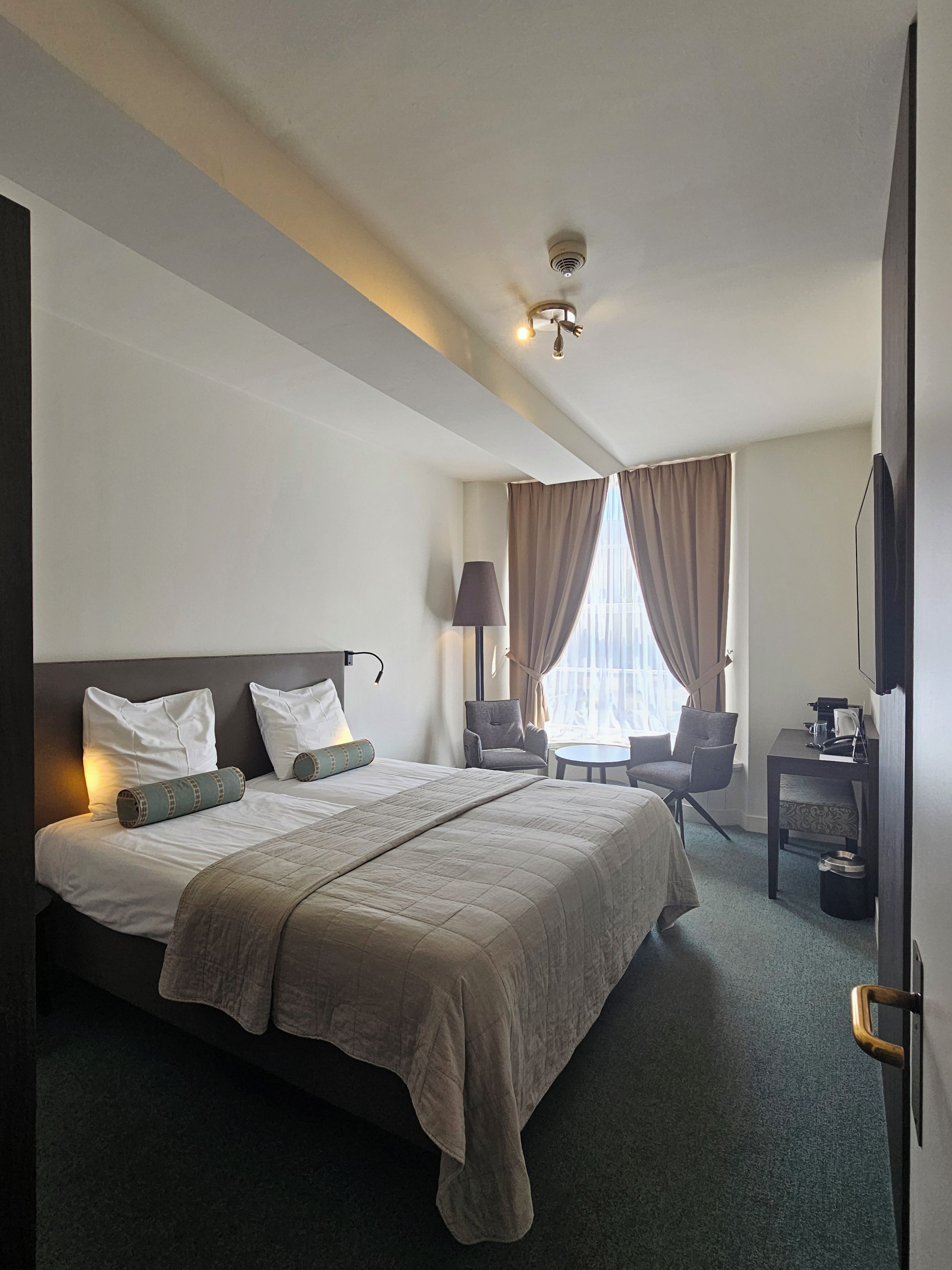 Ein Hotelzimmer mit einem großen Bett, einem Schreibtisch, einem Stuhl, einem Fernseher und einem Fenster mit Vorhängen.
