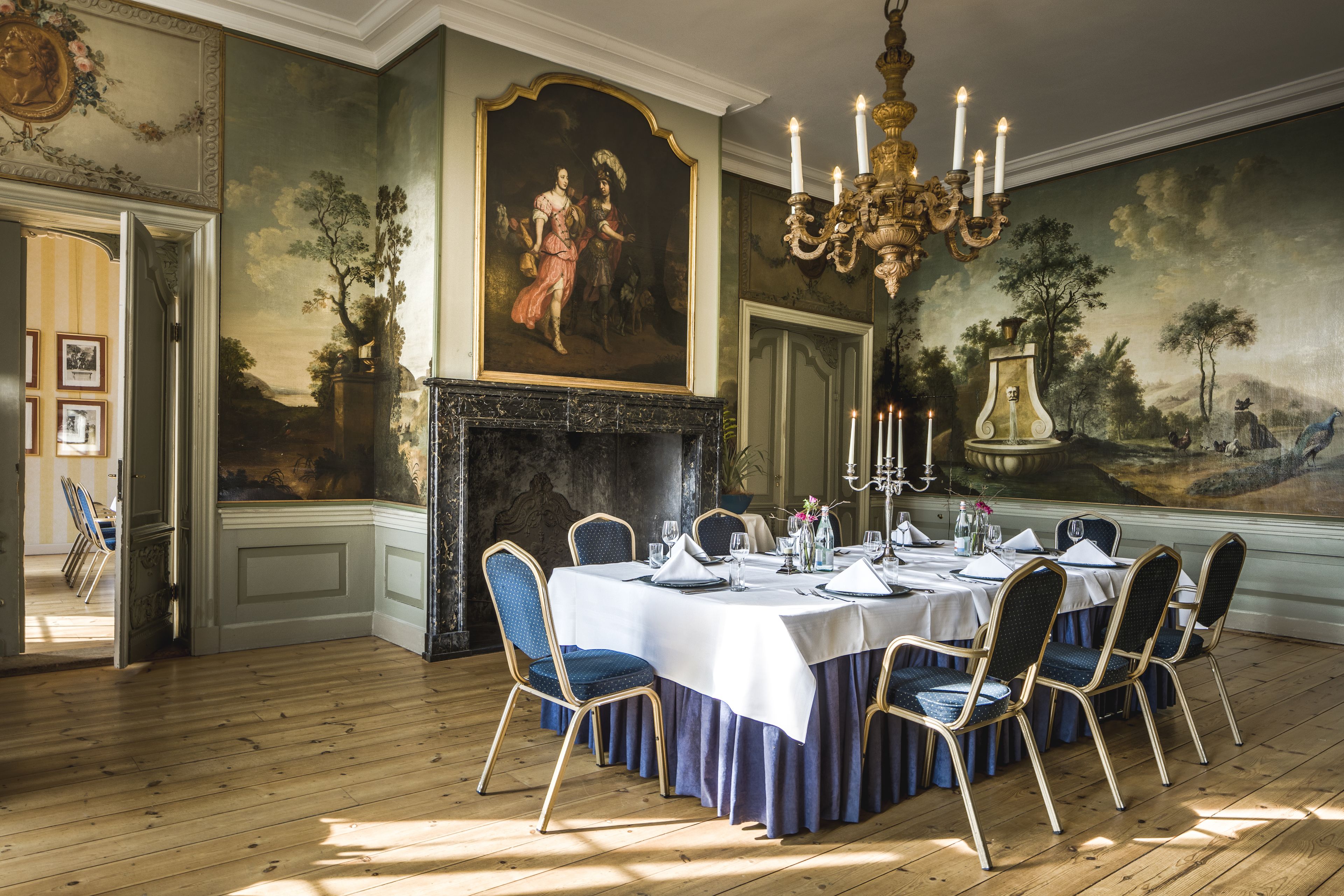 A formal dining room with a large table set for six, surrounded by upholstered chairs. A grand chandelier hangs above, and a fireplace with a mantelpiece is visible.