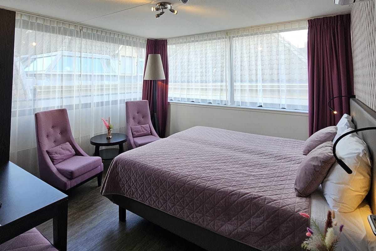Een hotelkamer met een groot bed, twee fauteuils, een klein tafeltje en een raam met gordijnen.