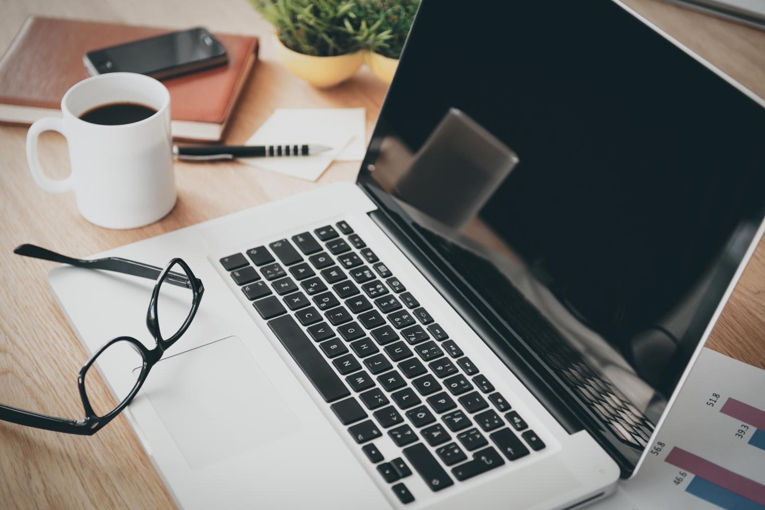Una computadora portátil sobre un escritorio con un par de lentes, una taza de café, un teléfono inteligente y una libreta.