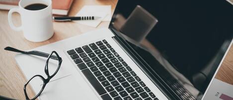 Una computadora portátil sobre un escritorio con un par de lentes, una taza de café, un teléfono inteligente y una libreta.