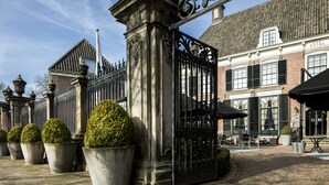 Een historisch gebouw met een hekpoort, grote beplante vormen en terrasplekken.