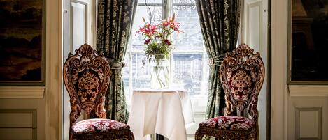 A room with two ornate chairs, a small table with a vase of flowers, and a window with patterned curtains.