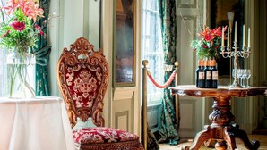 Een kamer met een houten vloer, een rode fauteuil, een tafel met een bloevas en een kandelaar met kaarsen.