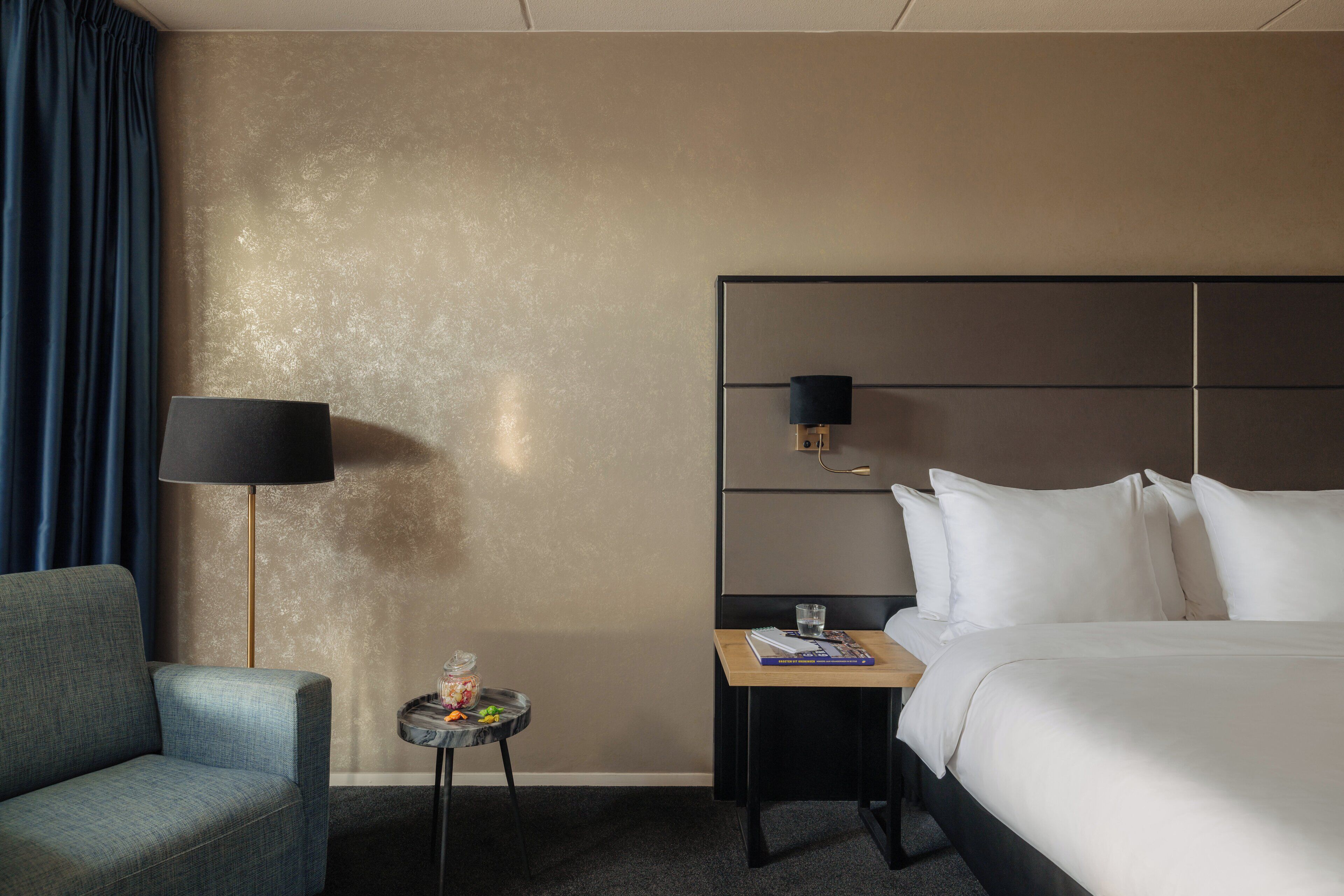 A hotel room with a bed, a nightstand, a lamp, a chair, and a small table with a glass and a book.