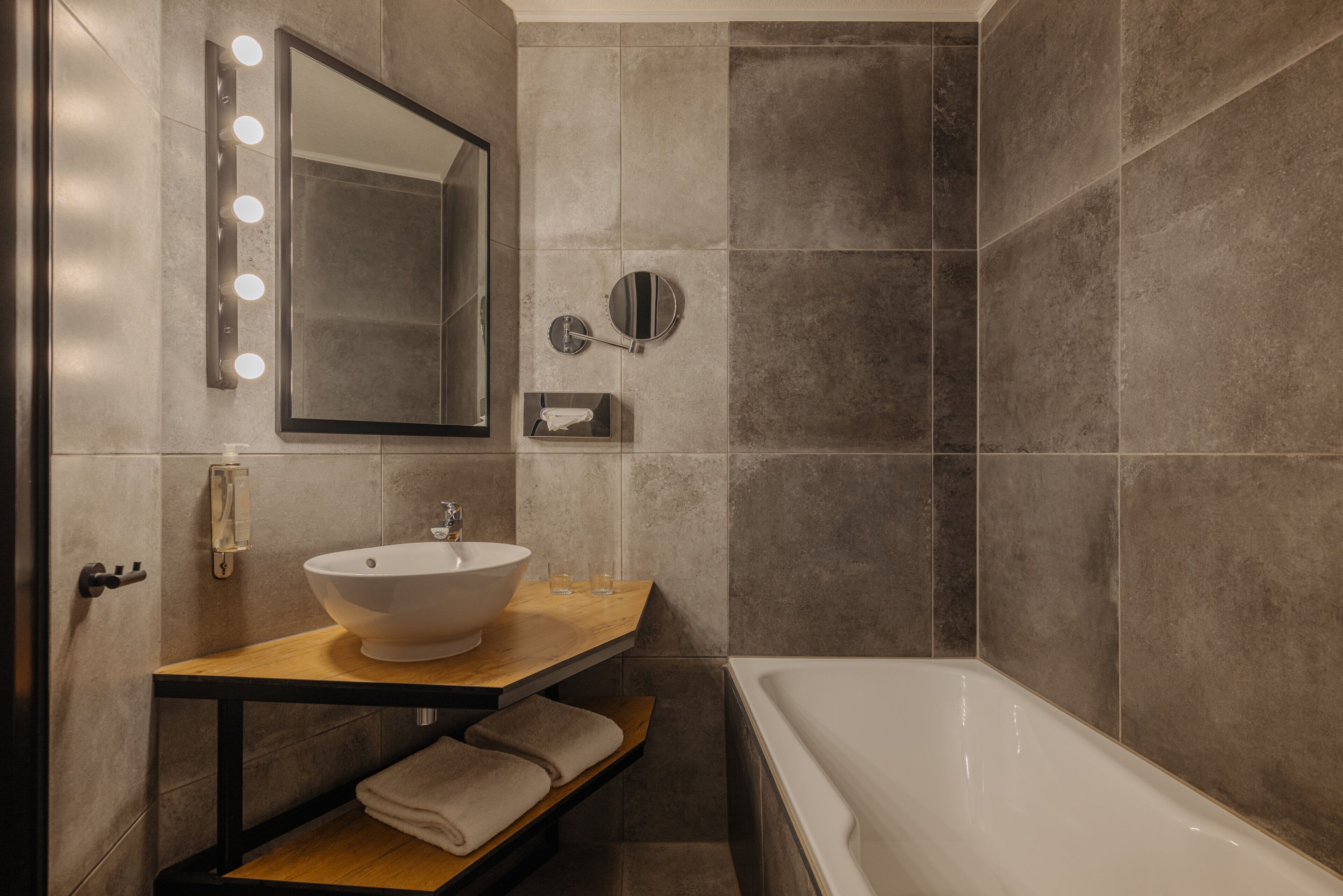 A modern bathroom with a large bathtub, a wooden vanity with a white sink, and a mirror with lights.