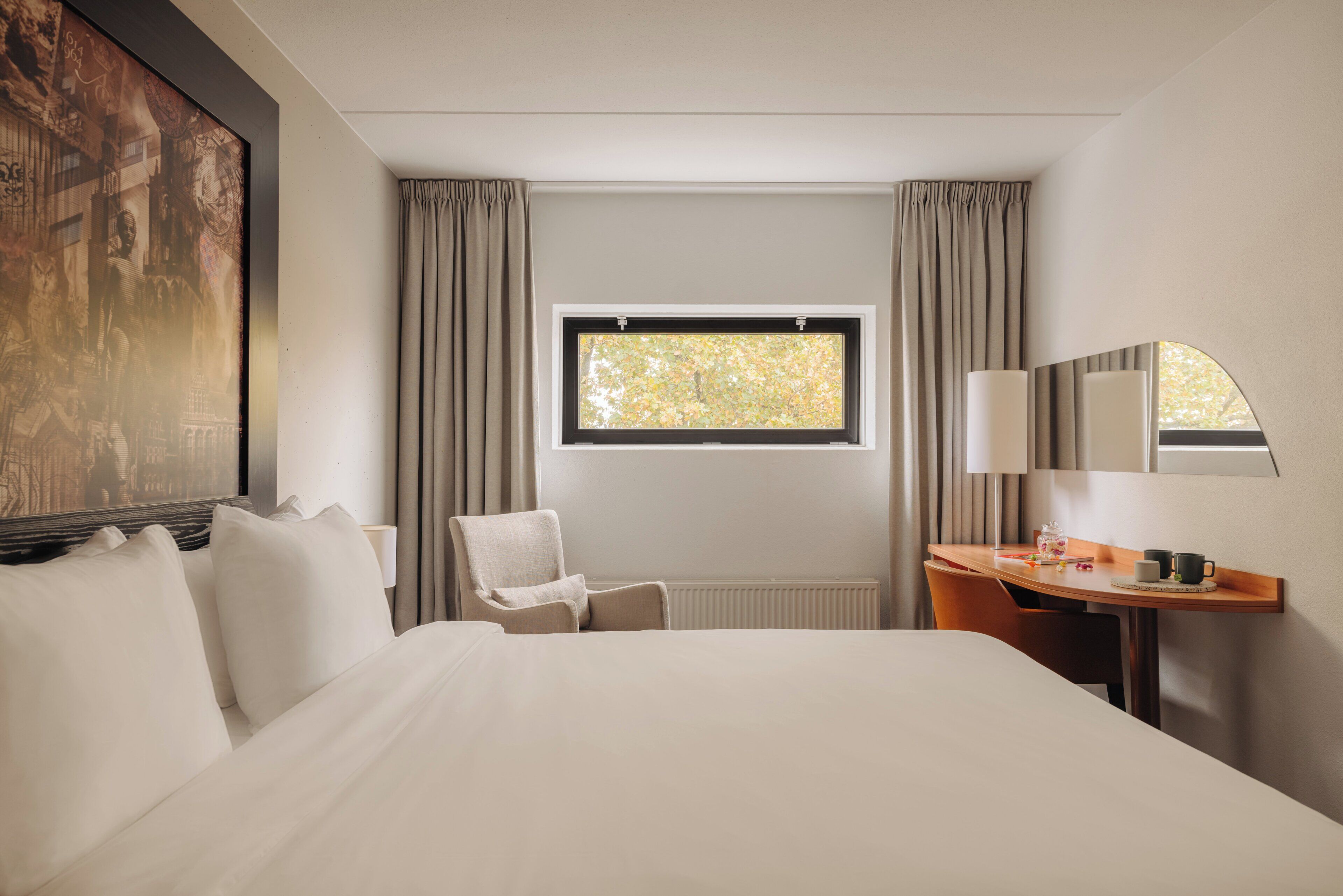 Une chambre d’hôtel avec un grand lit, un bureau avec une chaise, une fenêtre avec des rideaux et un tableau au mur.