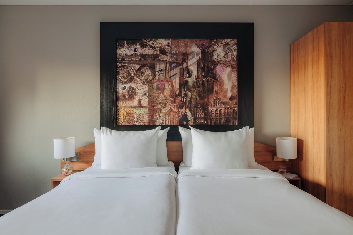 A double bed with white linens, two bedside tables with lamps, and a large abstract painting on the wall.