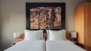 Una cama doble con ropa de cama blanca, dos mesitas de noche con lámparas y un cuadro abstracto grande en la pared.