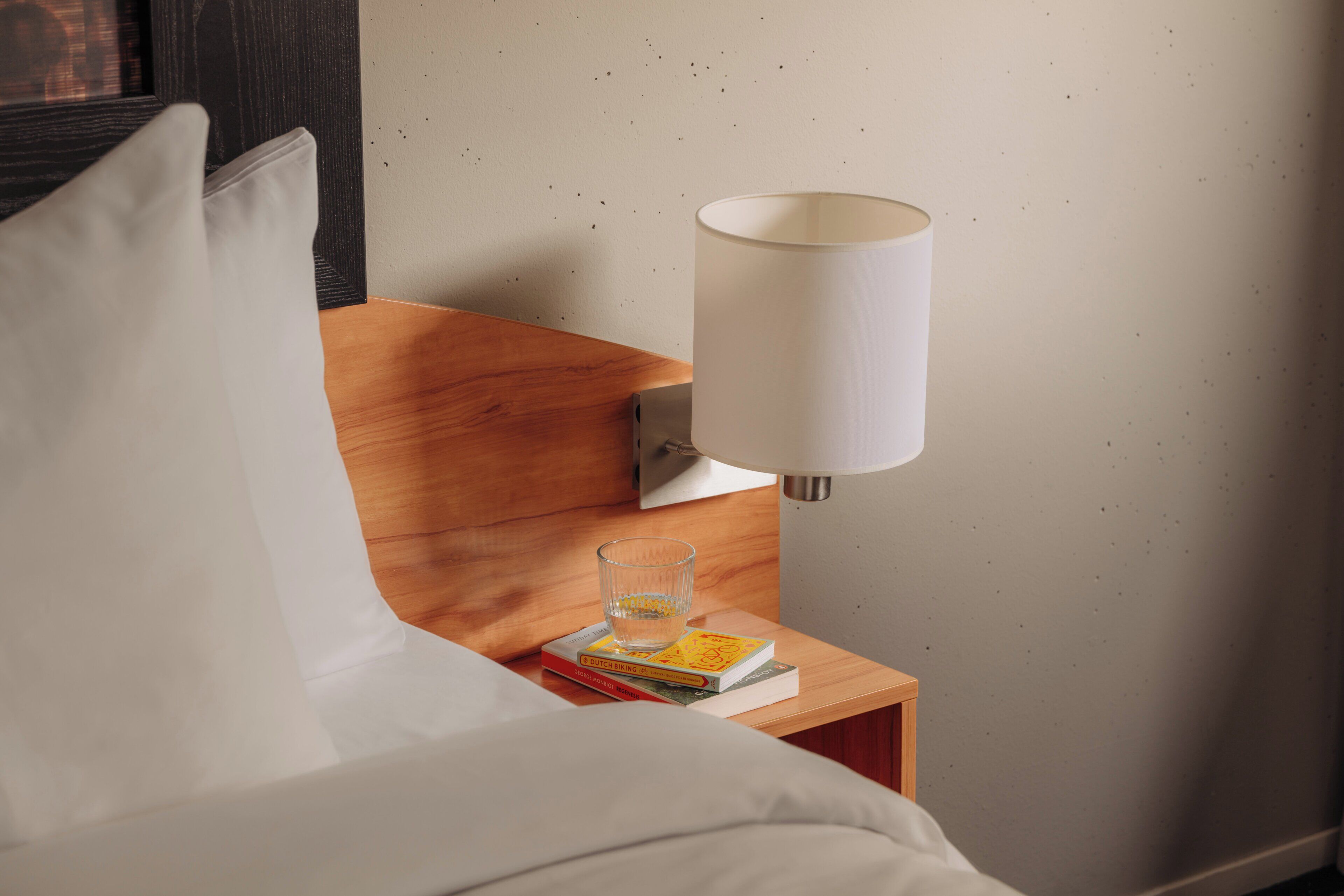 A neatly made bed with white sheets, a wooden nightstand with a lamp, and a glass of water.
