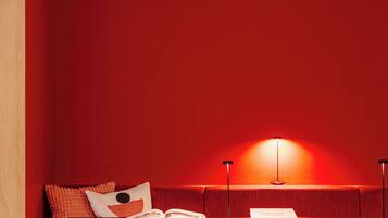 Een gezellig zitgedeelte met een rode bekleden bank, een kleine tafel met een boek en een lamp, en een houten wand.