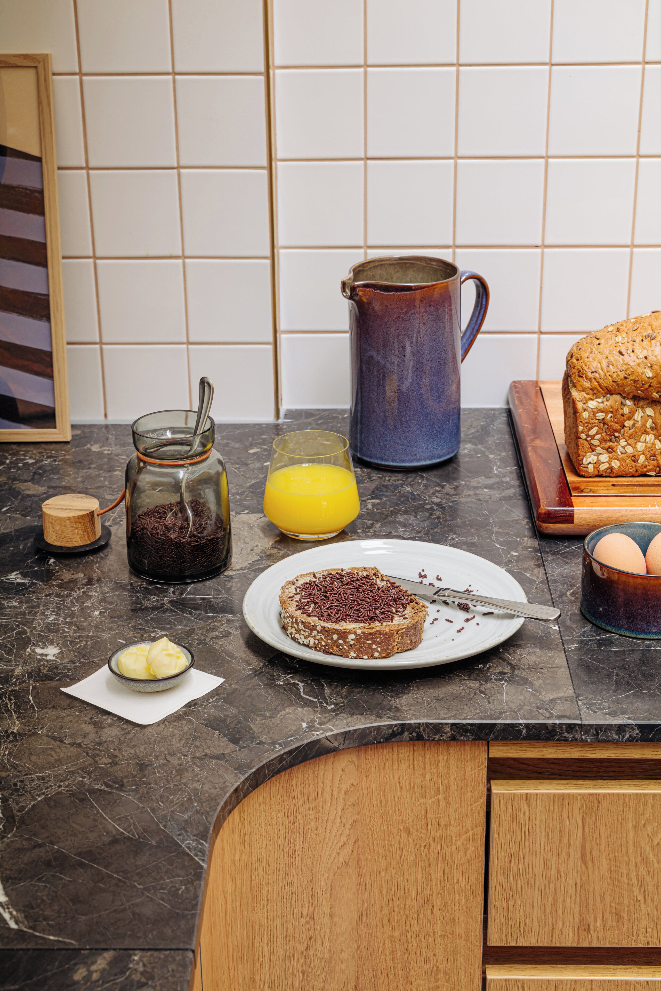 A kitchen counter with a coffee maker, a glass of orange juice, a loaf of bread, a plate with toast, and a bowl of sliced bananas.