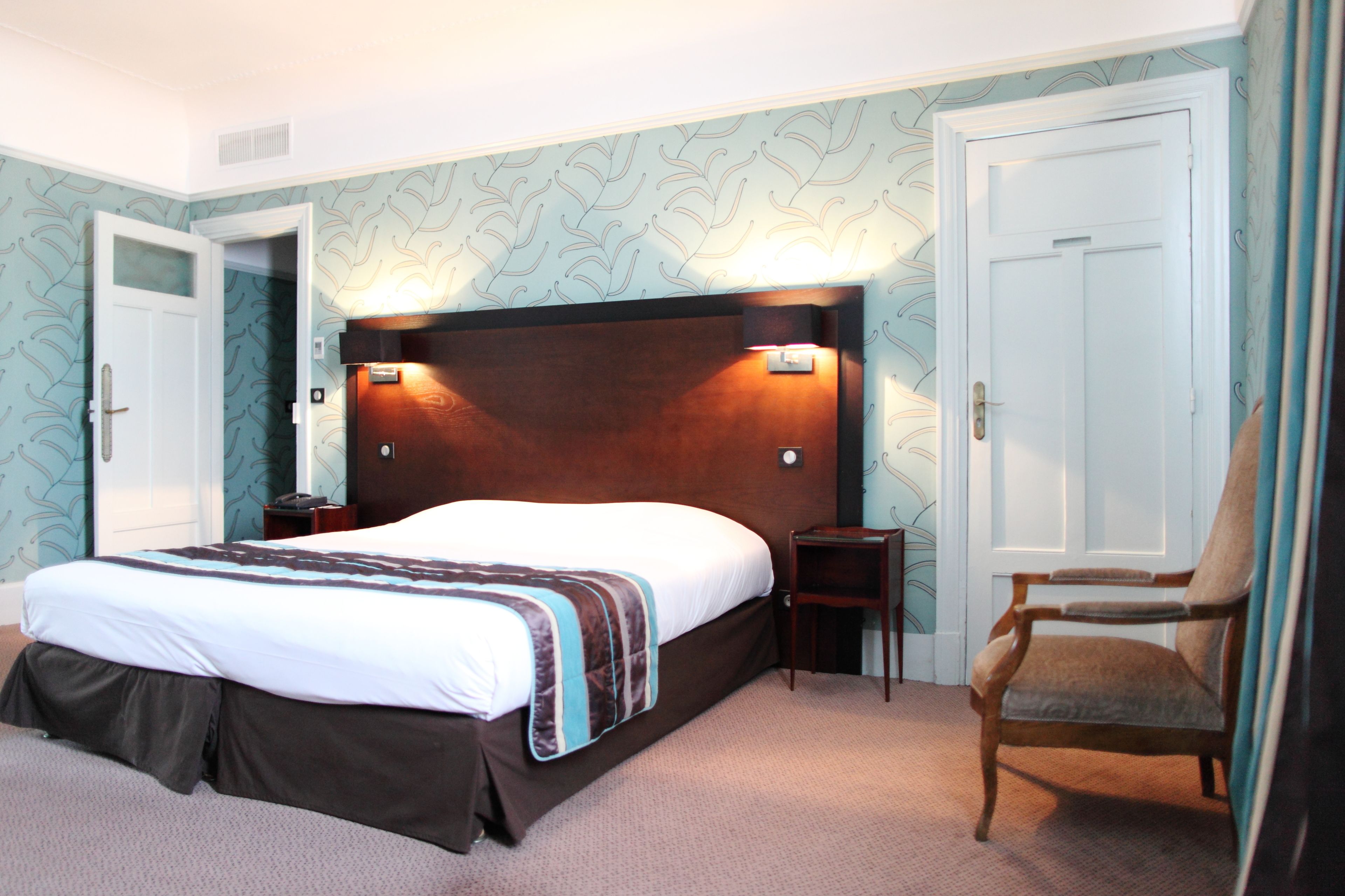 A hotel room with a large bed, two bedside tables, a wooden headboard, a chair, and patterned wallpaper.