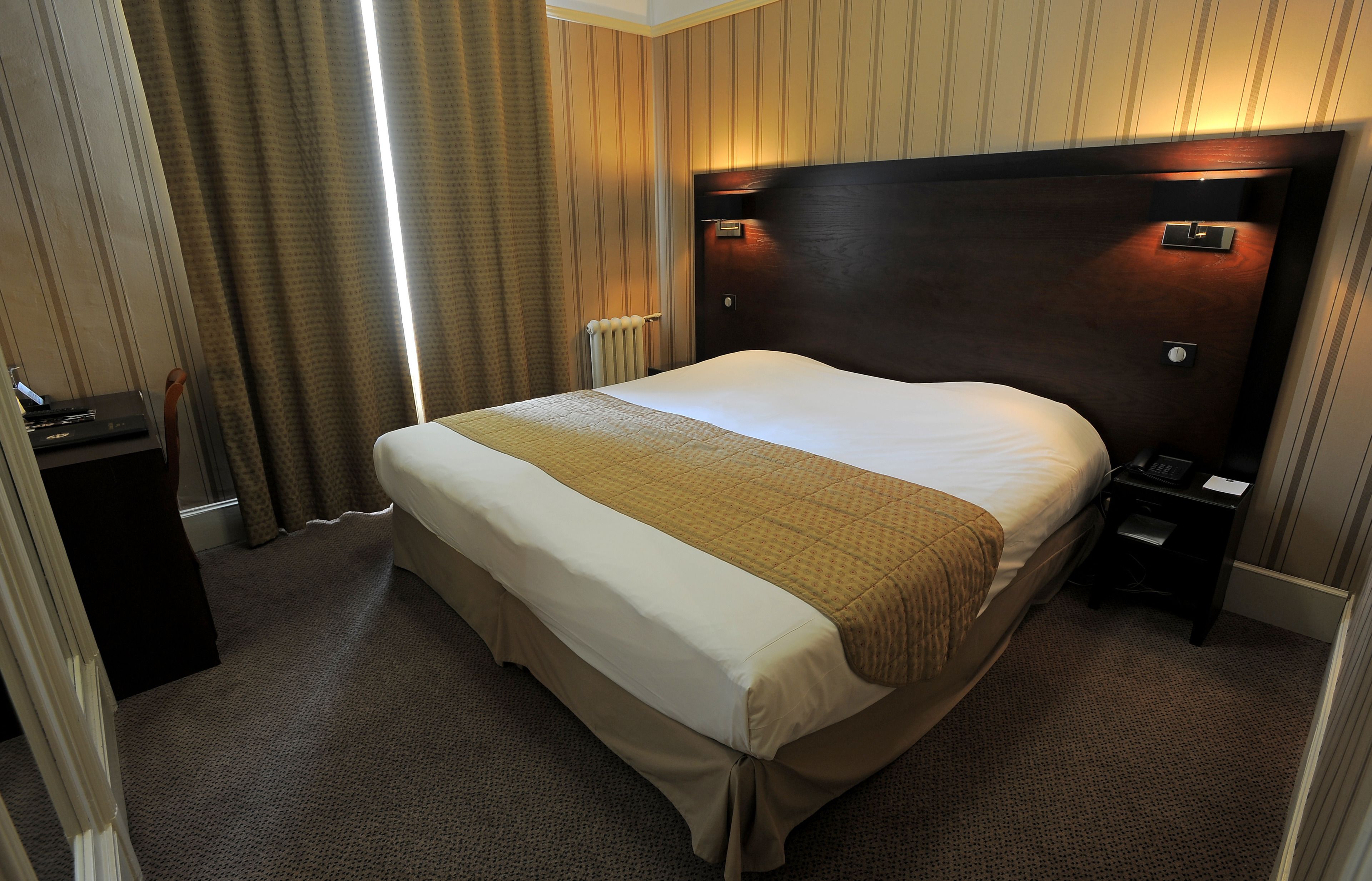 A hotel room with a large bed, a bedside table, a nightstand, a desk, and a radiator.