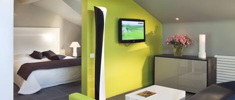 Una habitación de hotel moderna con una cama, un sillón verde, una mesita de centro blanca, un televisor montado en la pared y un jarrón con flores.