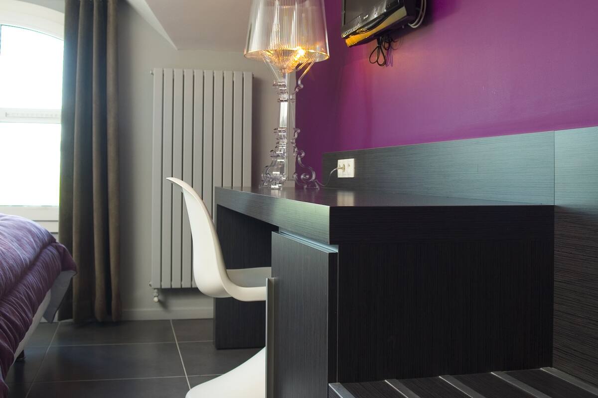 Ein modernes Hotelzimmer mit dunklem Holzschreibtisch, einem wandmontierten Fernseher, einem außergewöhnlichen weißen Sessel und einem großen Fenster mit Vorhängen.