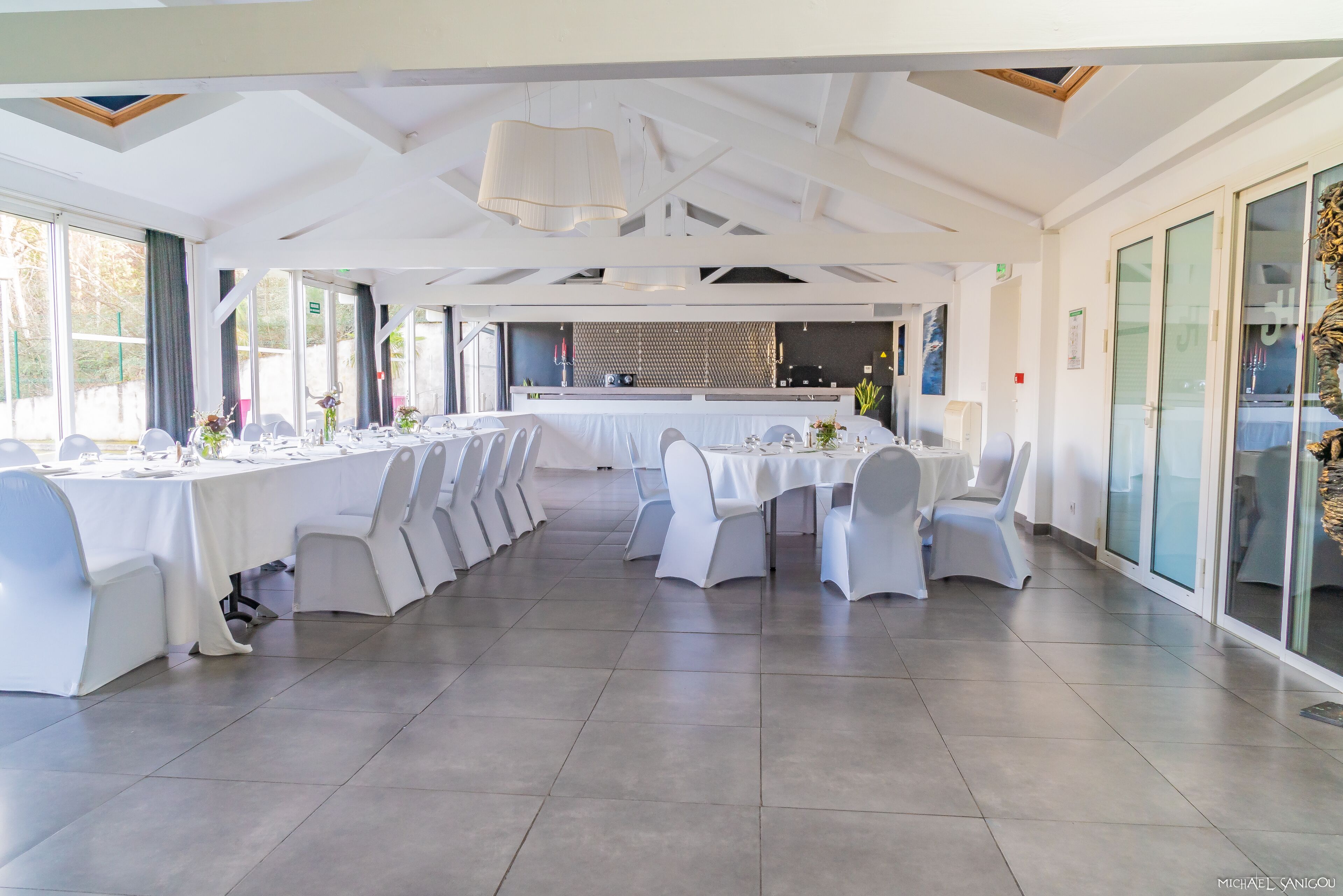 A spacious banquet hall with white tables and chairs, large windows, and a modern ceiling design.
