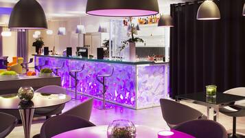 Un área de bar moderna con mostrador de vidrio, iluminación ambiental morada y diversas opciones de asientos, incluyendo taburetes y mesas redondas.