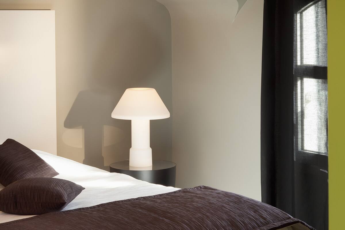 Ein ordentlich bezogenes Bett mit dunkler Bettwäsche, eine Nachttischlampe und ein Fenster mit Blick ins Freie.