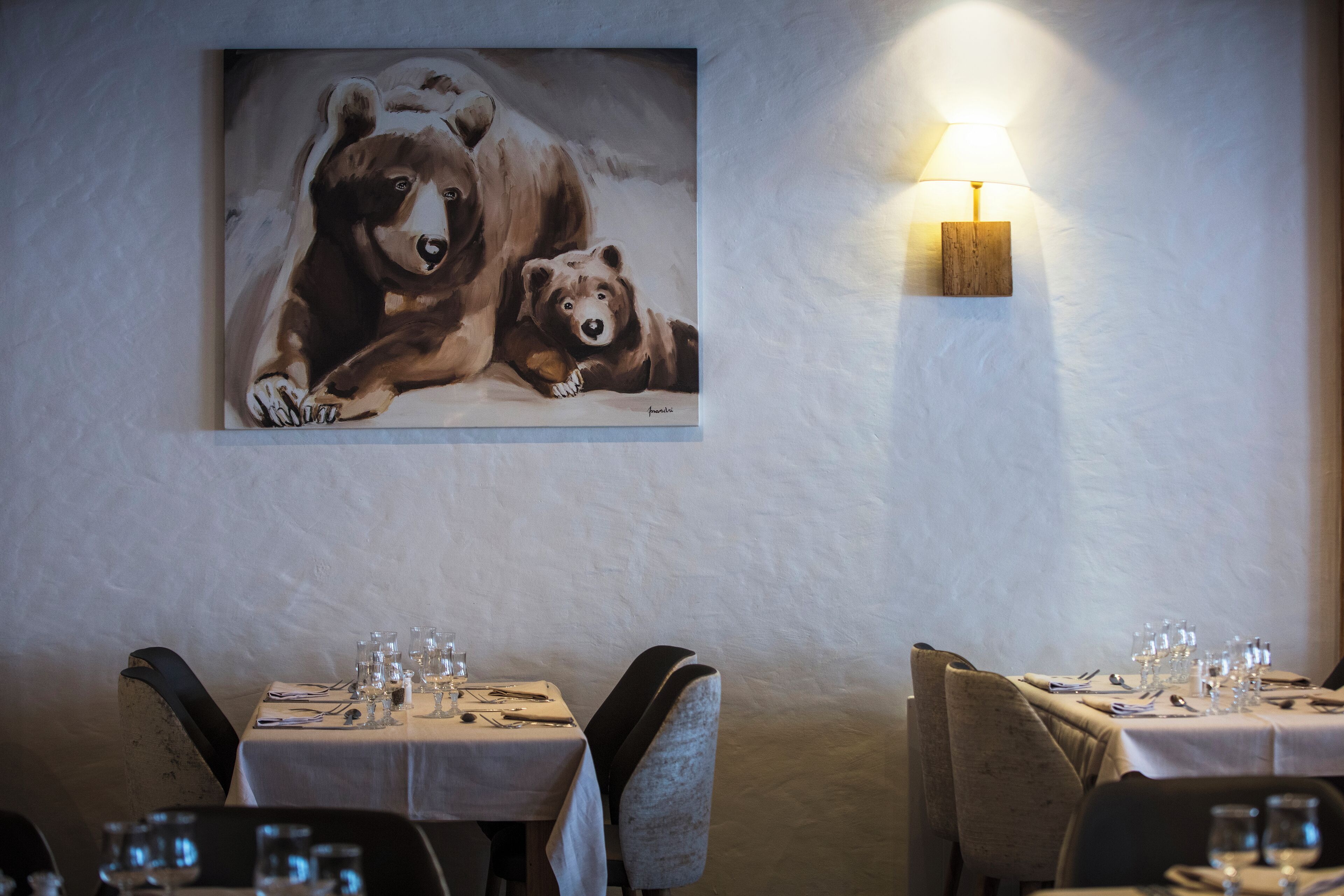 A restaurant with tables set for dining, a wall-mounted painting of bears, and a wall-mounted lamp.