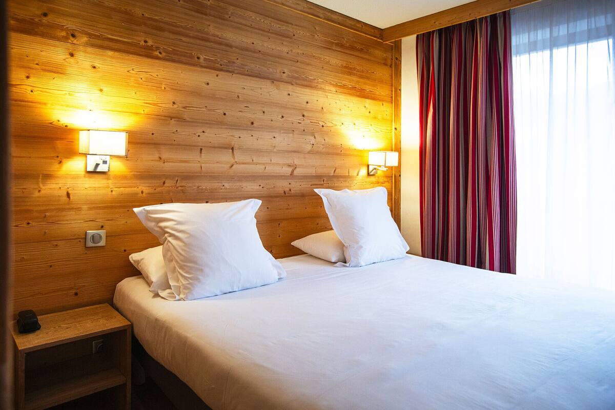 A hotel room with a wooden headboard, two white pillows, a bedside table with a lamp, and a window with striped curtains.