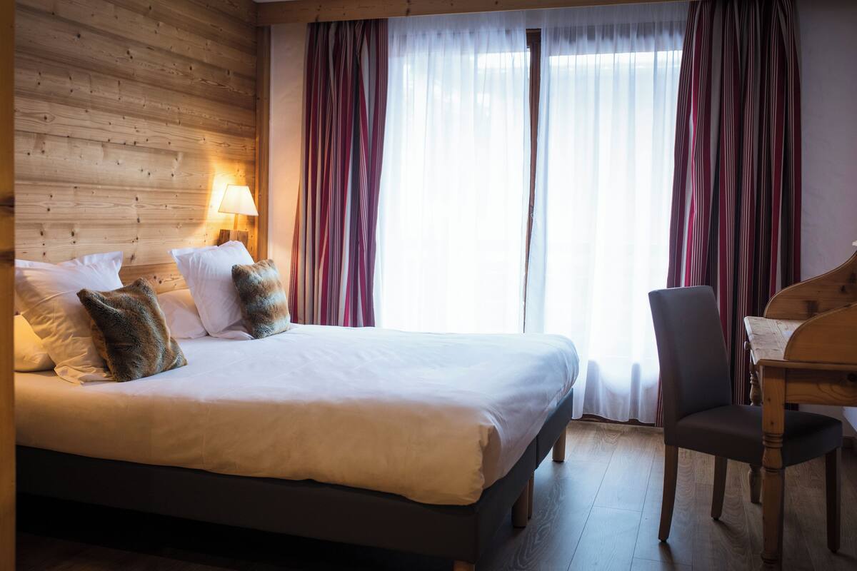 A neatly made bed with pillows, a wooden headboard, a chair, and a lamp in a room with wooden walls and a large window with curtains.