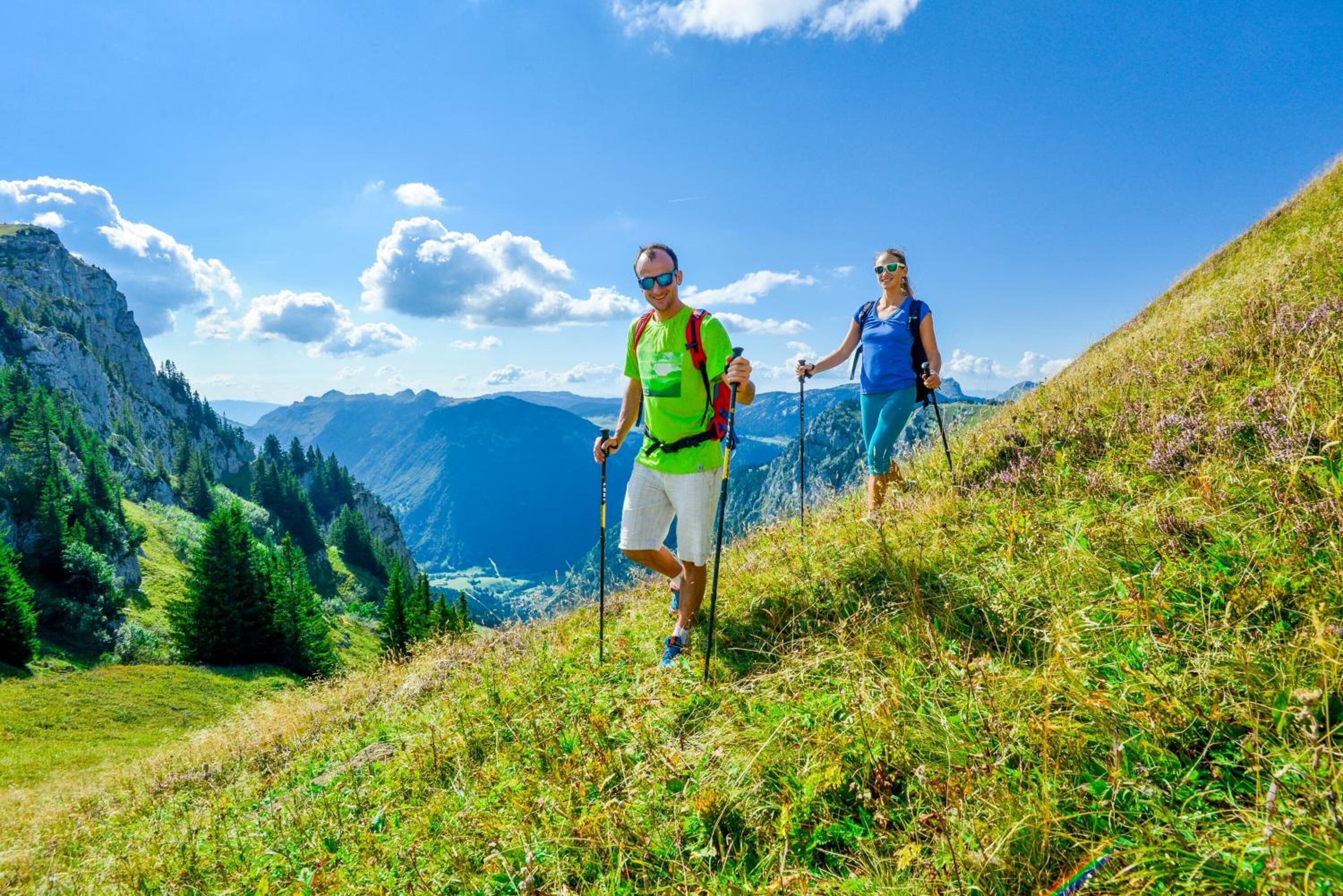 Two people hiking on a grassy hillside with mountains in the background.