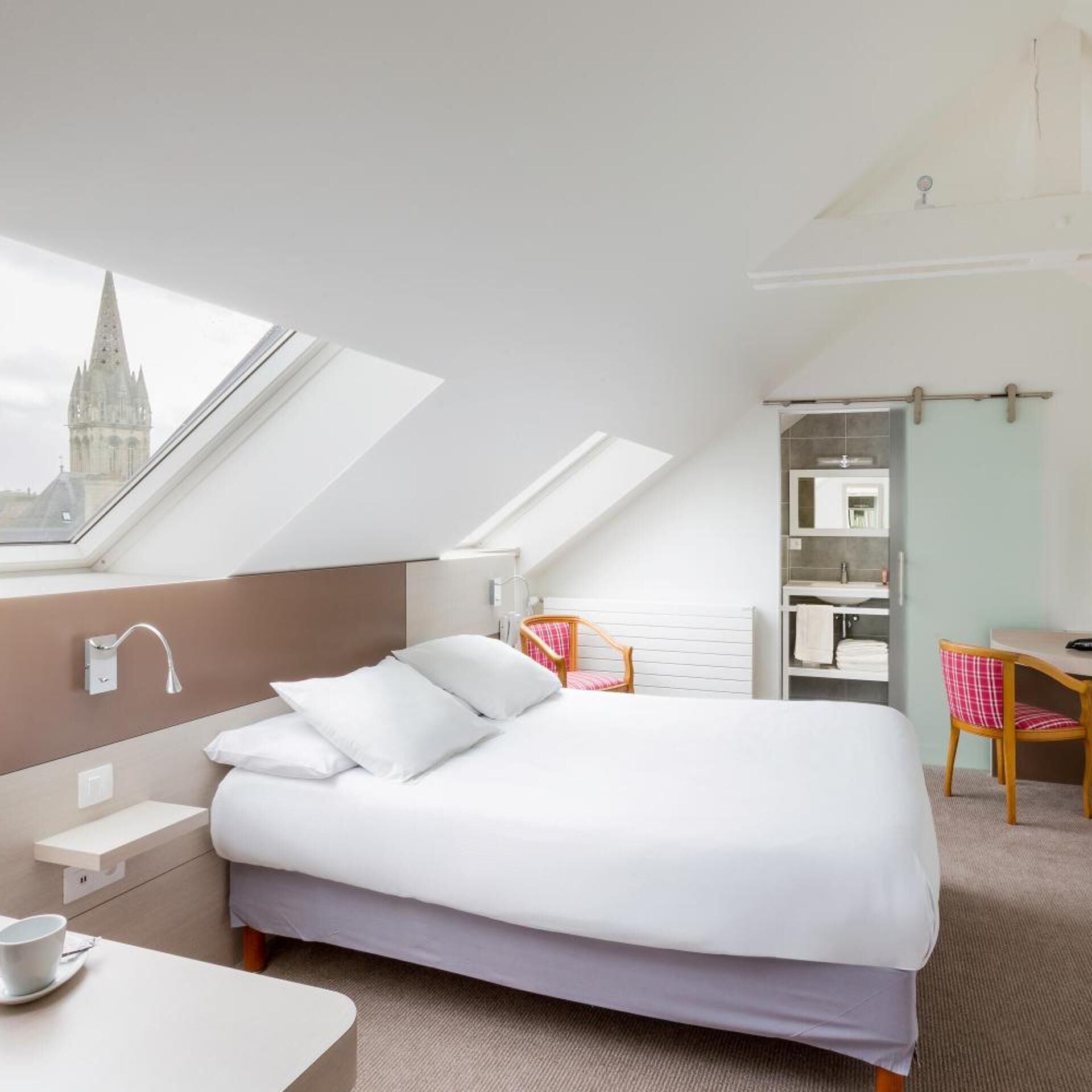 A hotel room with a bed, a desk, a chair, a bathroom, and a skylight.