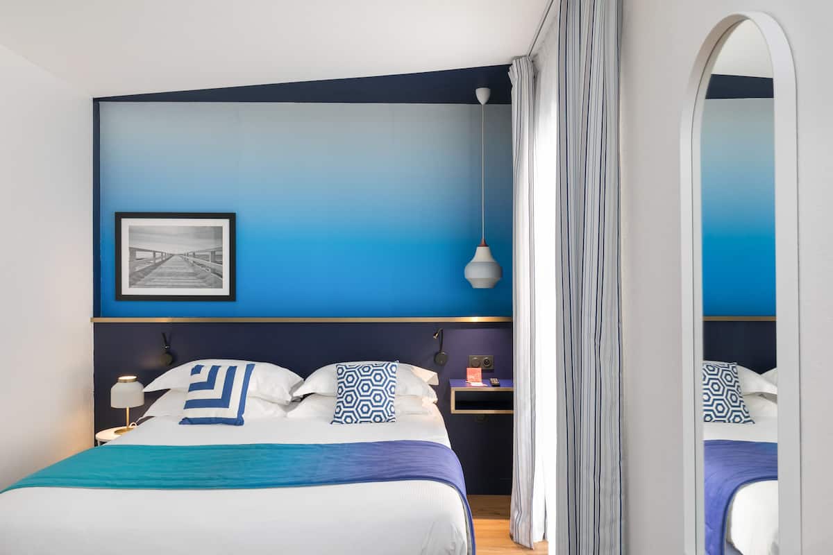 A modern hotel room with a large bed, a bedside table, a nightstand, a framed picture on the wall, and a mirror.