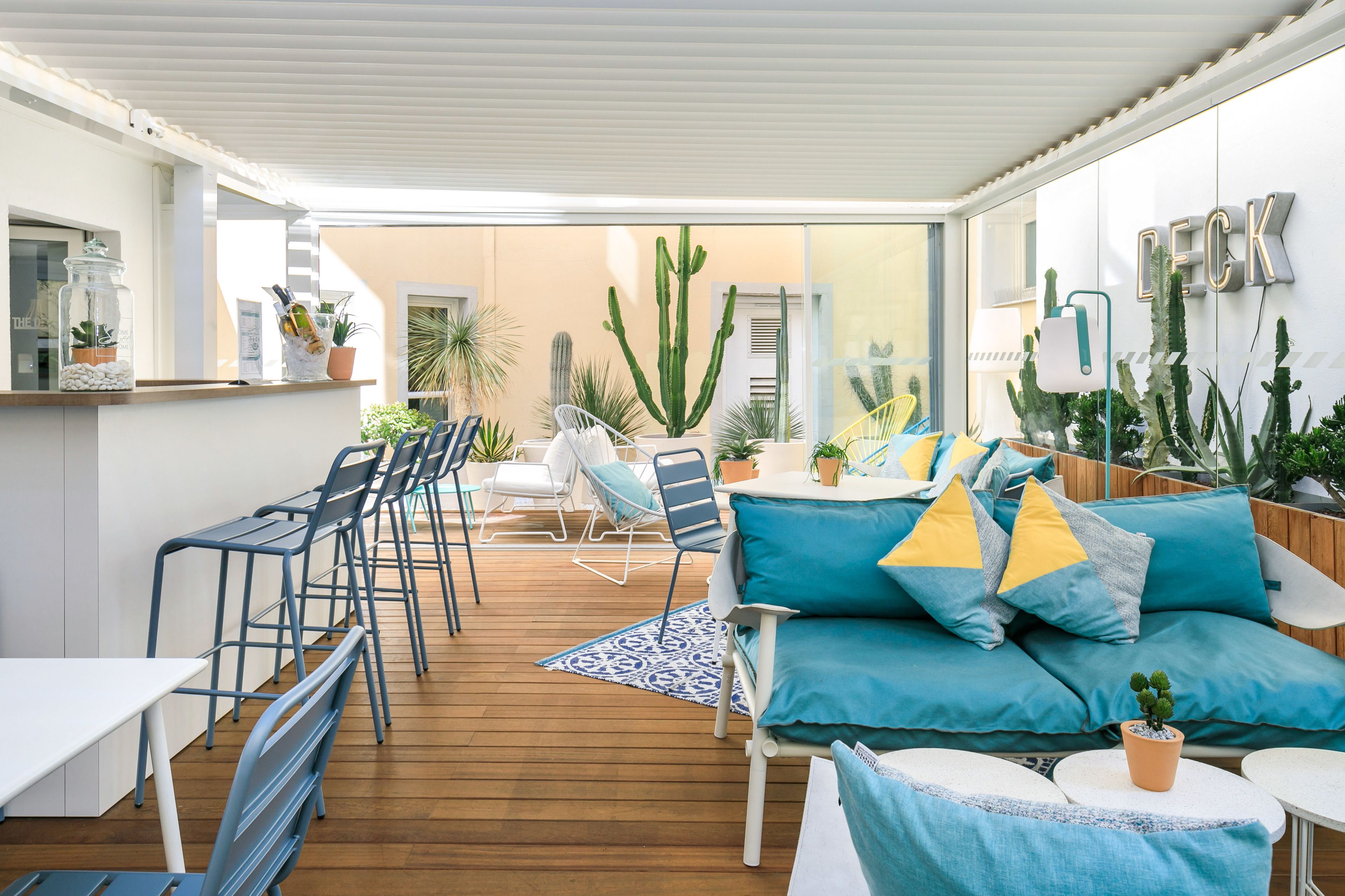 Un patio exterior con suelo de madera, muebles azules y blancos, y diversas macetas con plantas.