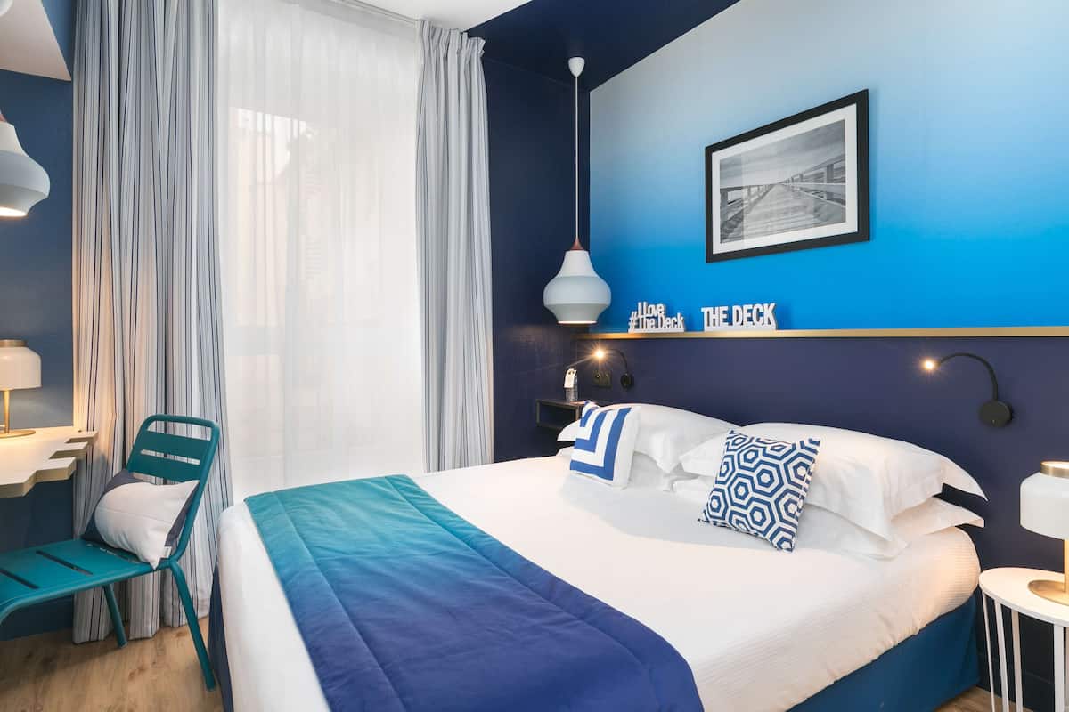 A modern hotel room with a large bed, a desk, and a chair. There is a window with sheer curtains, a wall-mounted picture, and a sign that reads "Loo # the Deck".
