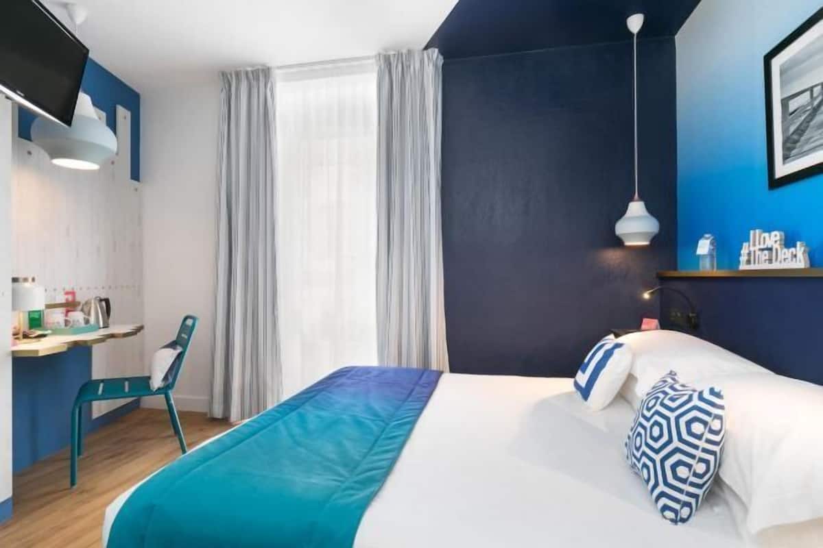 A hotel room with a bed, a desk, a chair, a window with curtains, and a wall-mounted TV.