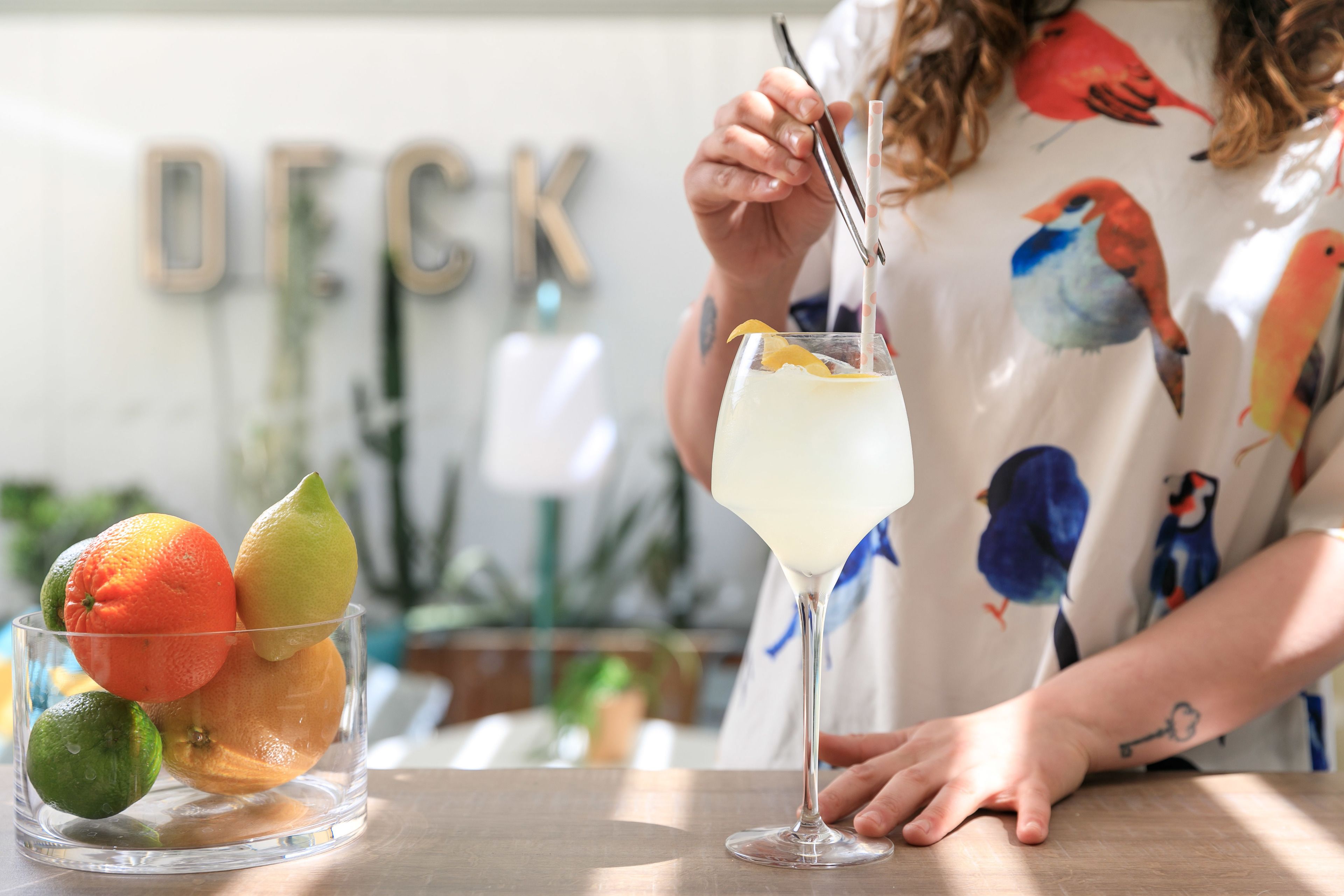 A person holding a cocktail glass with a straw, standing next to a bowl of fruit.