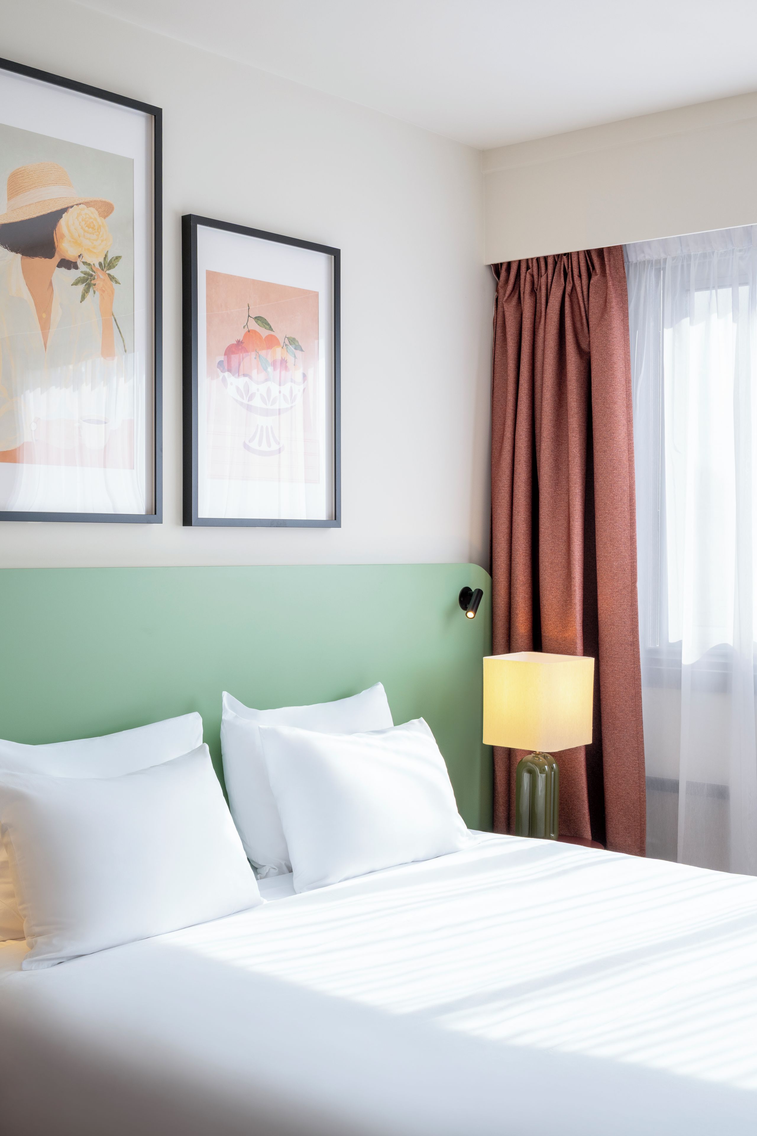 A hotel room with a bed, two framed artworks, a lamp, and a window with curtains.