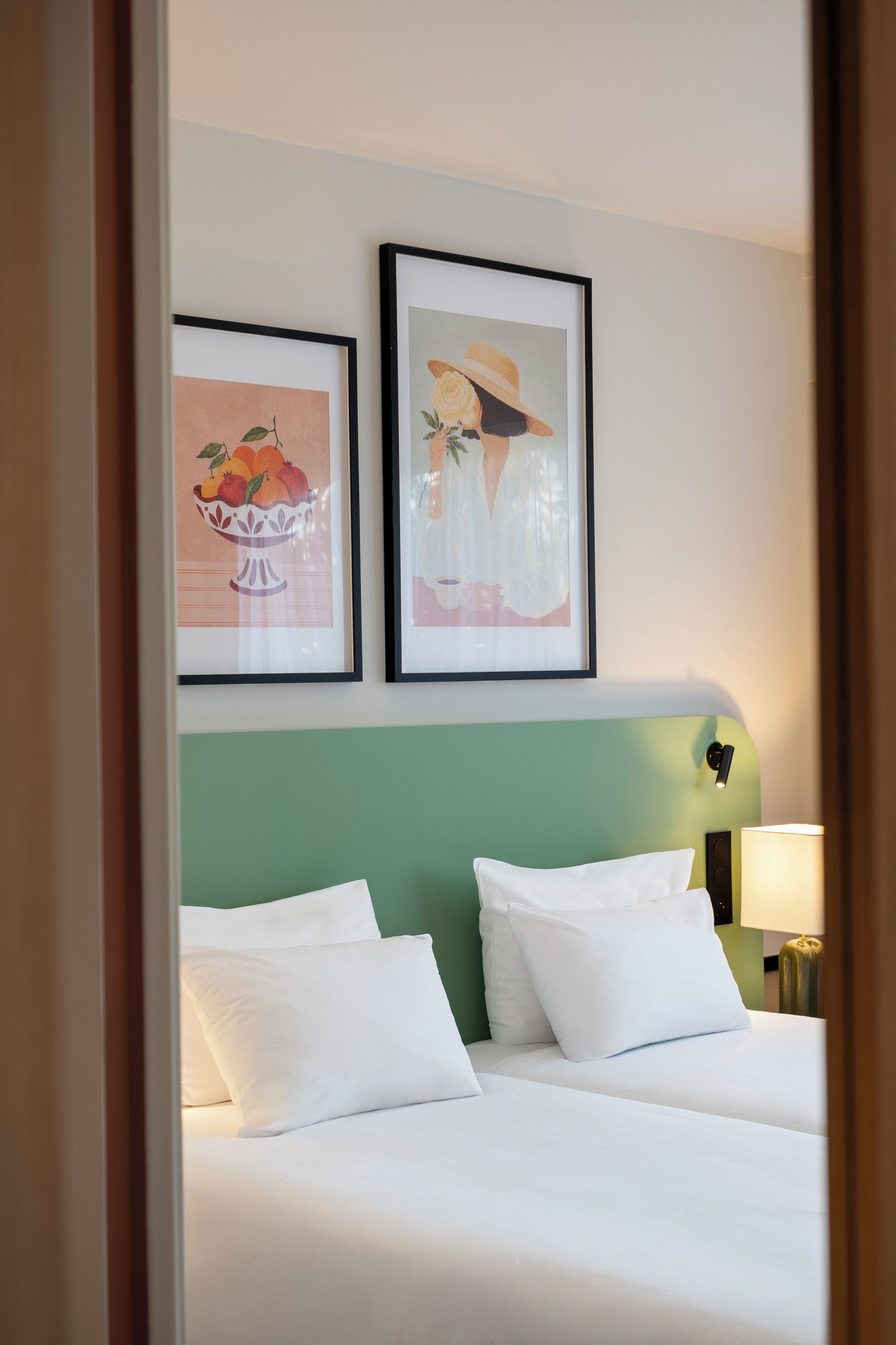 Ein Hotelzimmer mit zwei Betten, einem grünen Kopfteil und zwei gerahmten Kunstwerken an der Wand.