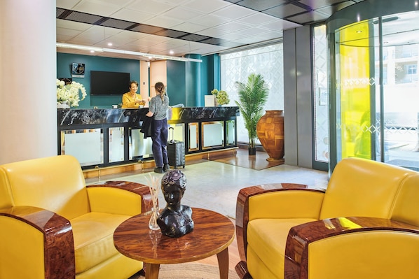 Un área de recepción con mostrador, dos recepcionistas, sillas amarillas, una mesa de madera con una escultura y una planta grande en maceta.