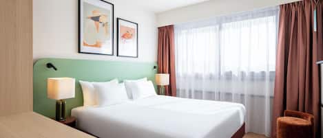 A hotel room with a large bed, two bedside tables, a desk, and a chair. There are two framed pictures on the wall.