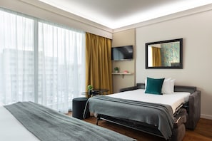 Habitación de hotel moderna con una cama grande, un televisor de pantalla plana, una mesita con una planta y una ventana con cortinas translúcidas.