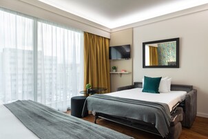 Habitación de hotel moderna con una cama grande, un televisor de pantalla plana, una mesita con una planta y una ventana con cortinas translúcidas.