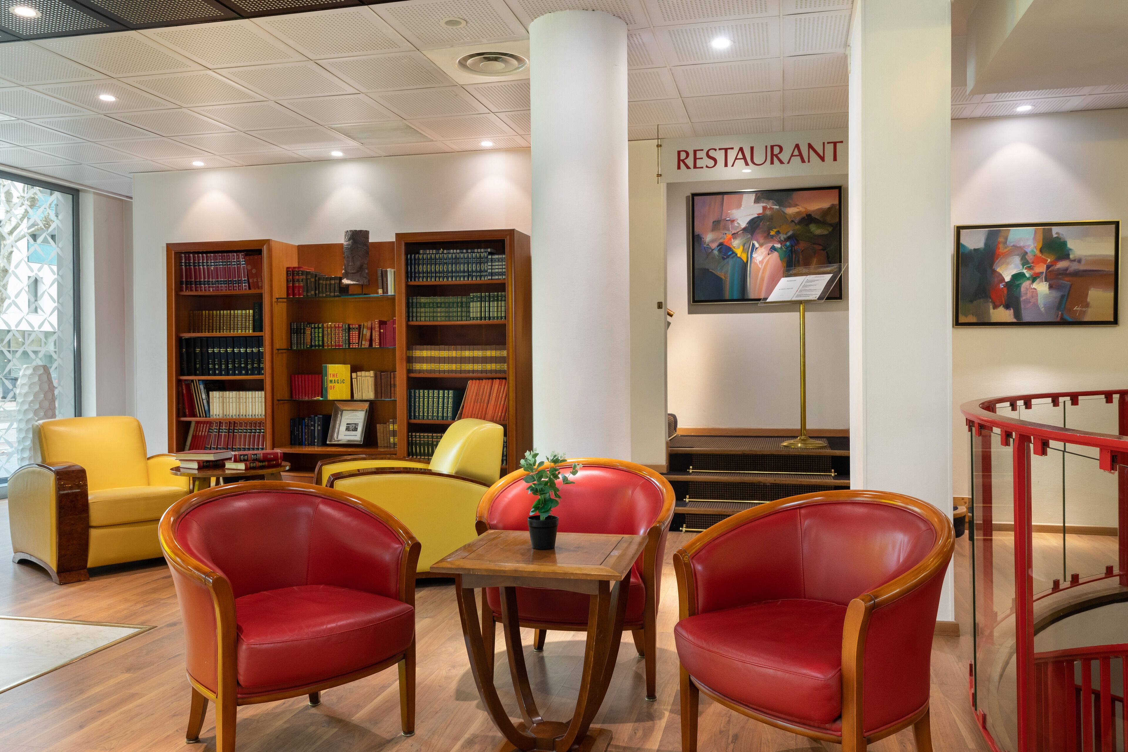 Une bibliothèque avec des étagères, un fauteuil rouge et jaune, une petite table avec une plante, et un panneau indiquant un restaurant.