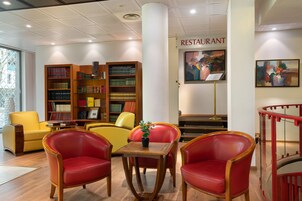 Una biblioteca con scaffali, una poltrona rossa e gialla, un tavolino con una pianta e un cartello che indica un ristorante.