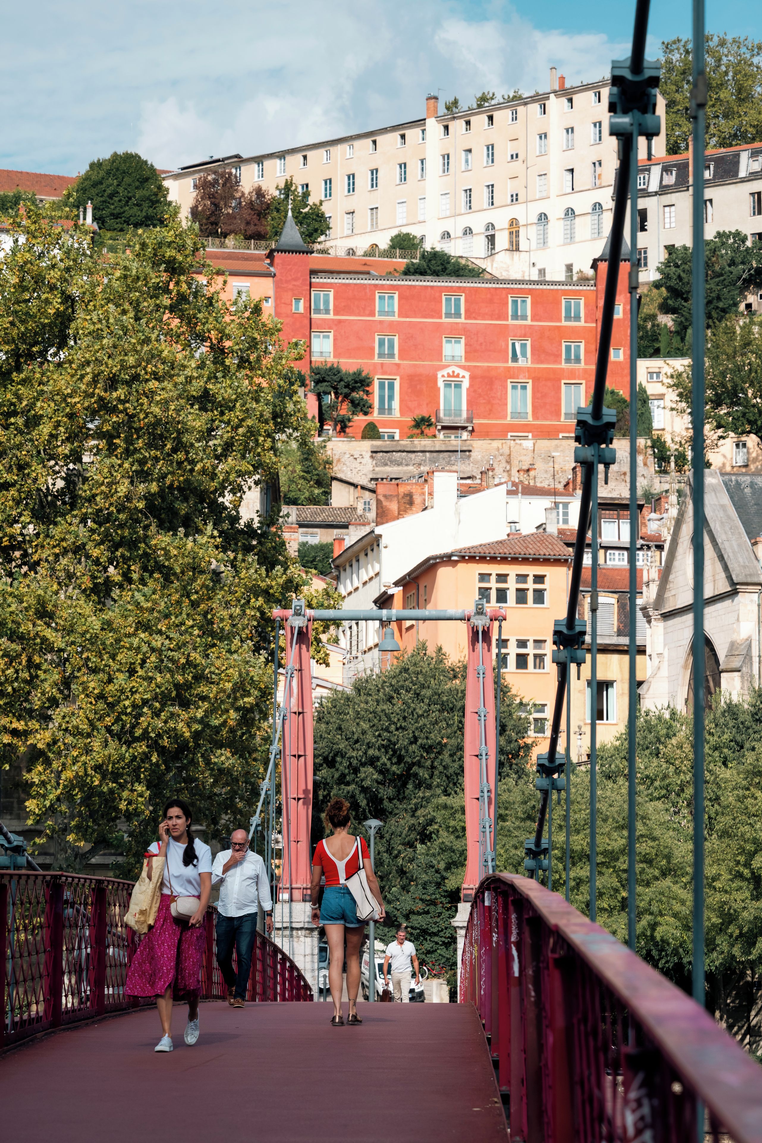 Un puente peatonal con barandilla roja, con vistas a un paisaje urbano con edificios y árboles.