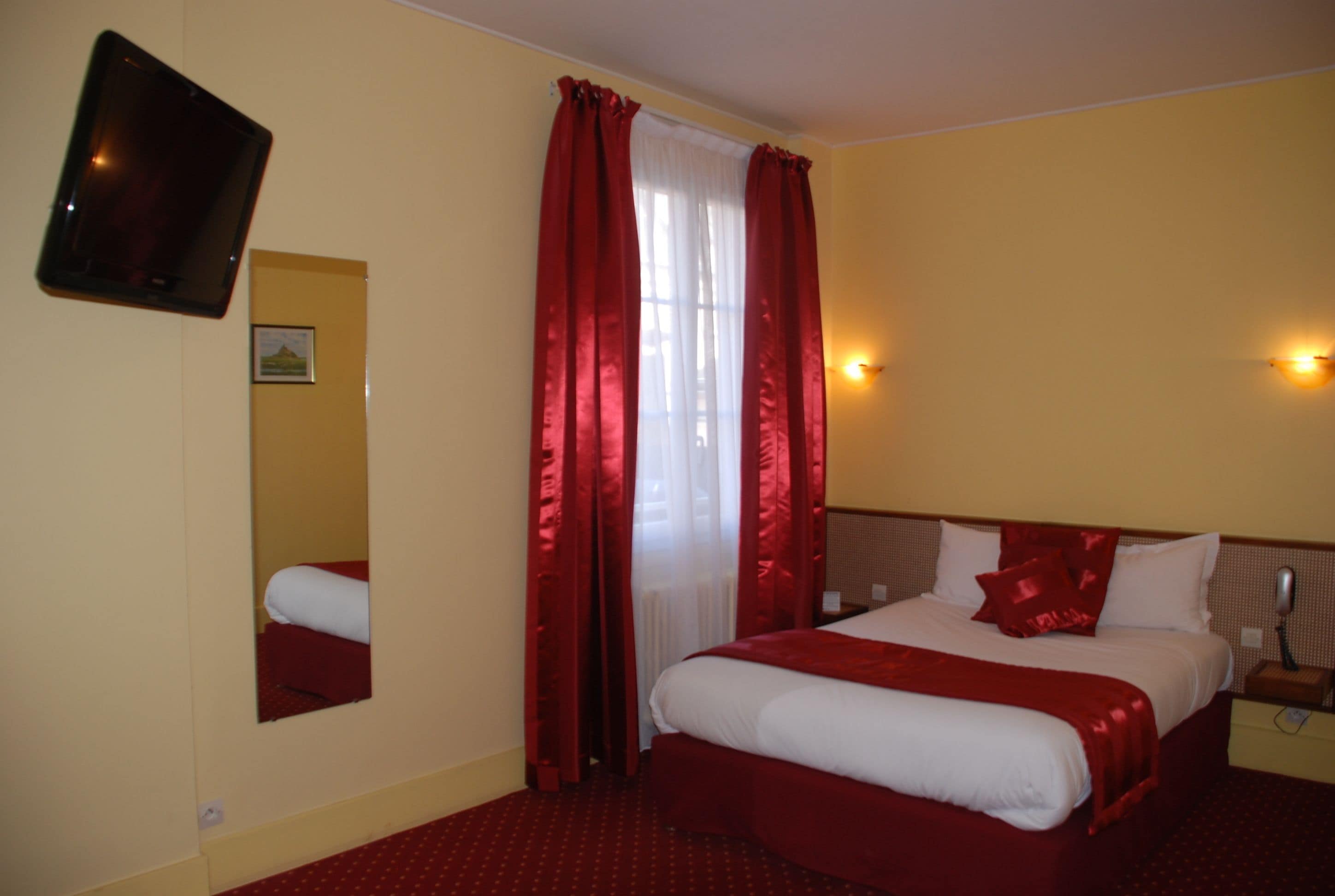 A hotel room with a bed, a TV mounted on the wall, a mirror, and a window with red curtains.