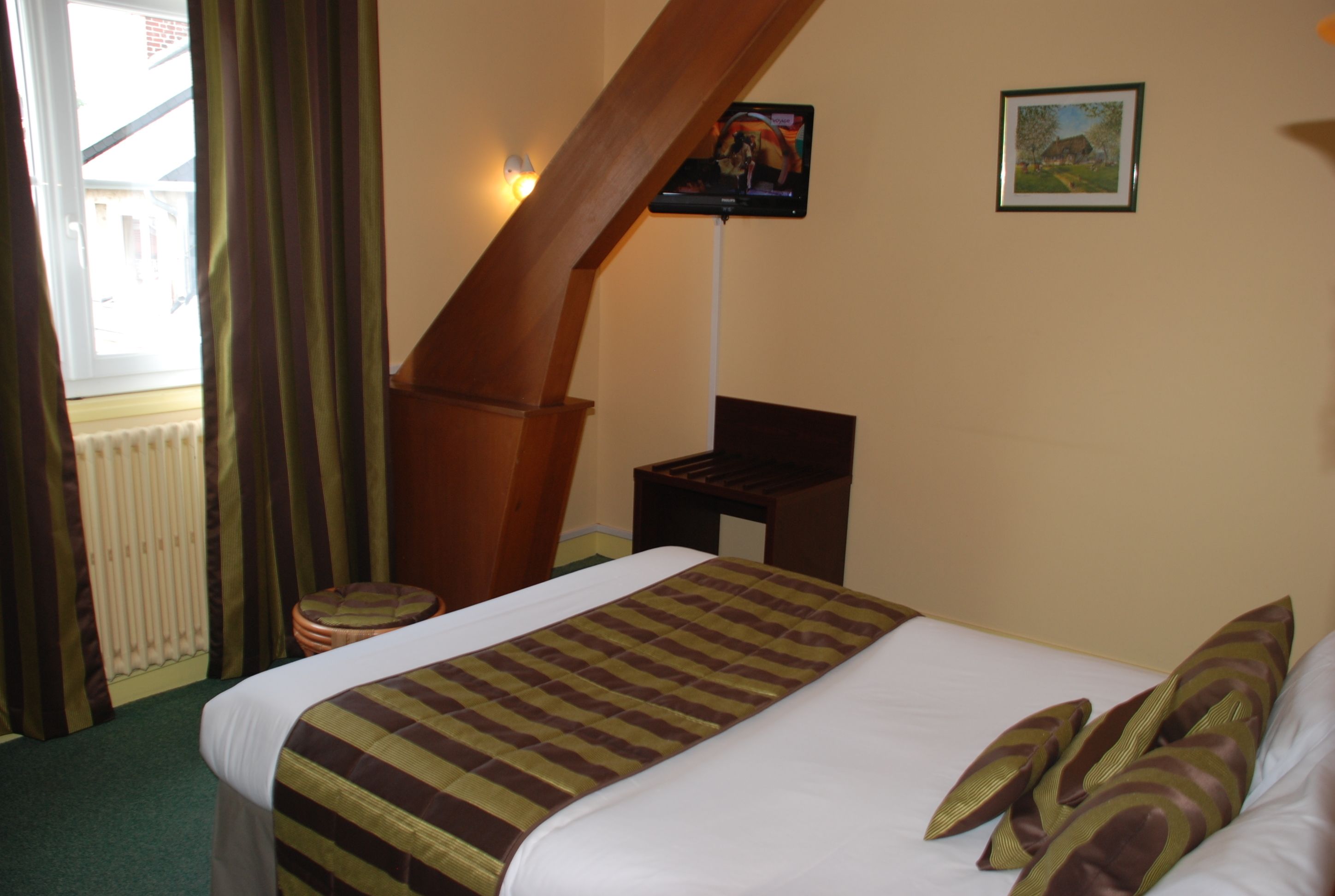 A hotel room with a bed, a television on a stand, a framed picture on the wall, and a window with curtains.