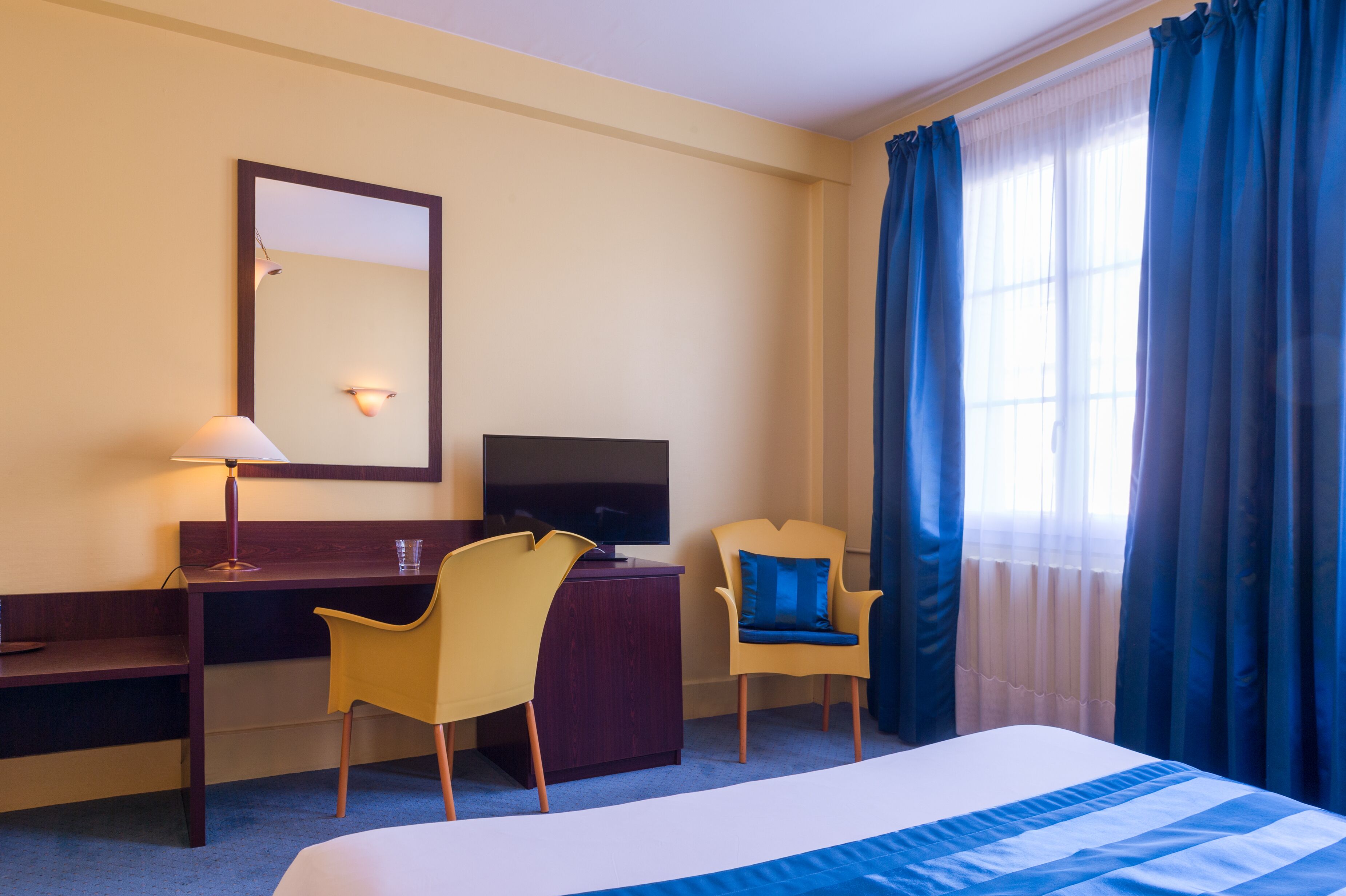A hotel room with a bed, a desk, two chairs, a TV, and a large window with curtains.