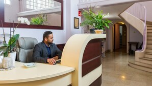 Un homme est assis à un bureau dans le hall d’un hôtel, devant un grand miroir, avec des plantes en pot et un escalier à proximité.