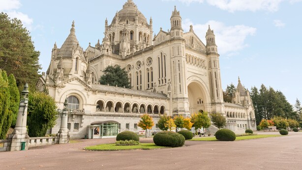 Un imposant bâtiment historique, doté de plusieurs dômes et d’arcades, entouré de jardins bien entretenus et d’une allée pavée.