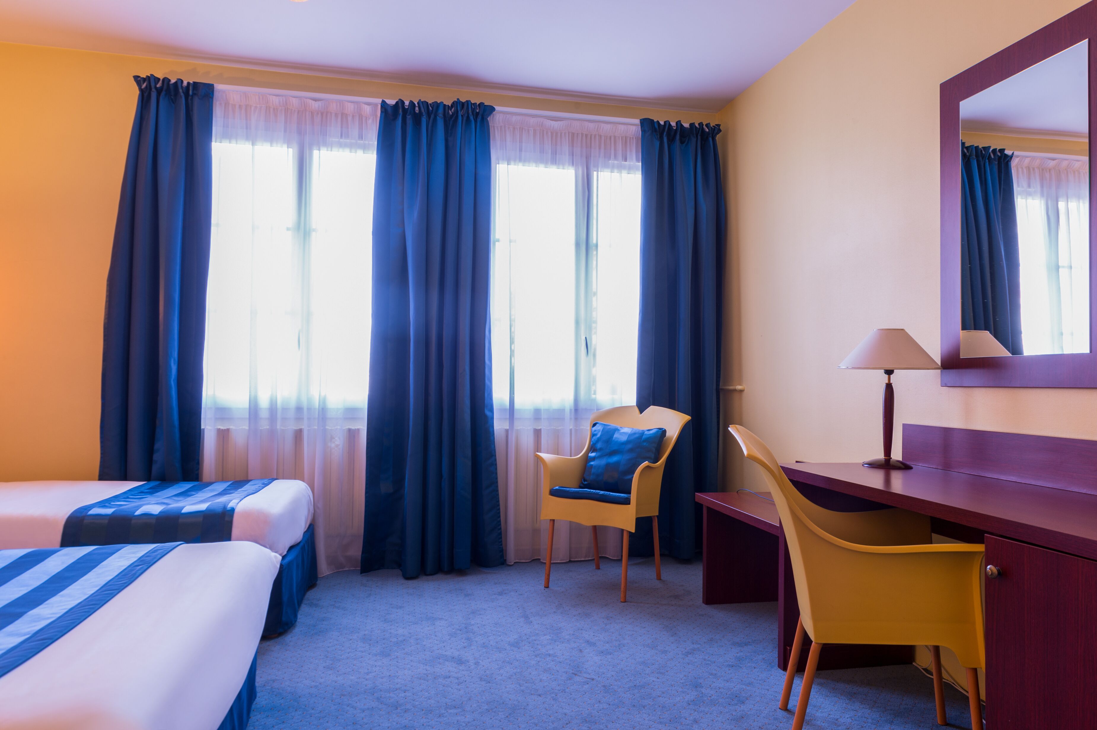 A hotel room with two beds, a desk, a chair, and a large window with curtains.
