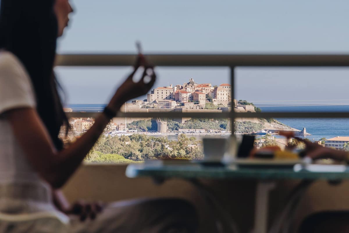 A person seated at a table with a view of a coastal town and buildings in the distance.